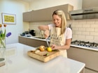 A smiling woman holding a wooden cutting board in a cozy kitchen setting.
