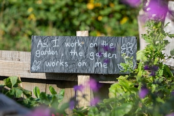 A wooden fence supports a dark slate sign with white chalk writing that reads, 'As I work on the garden, the garden works on me,' accompanied by simple drawings of a flower and a bee. Lush green plants surround the sign, and purple flowers are visible in the foreground with a slightly blurred effect.
