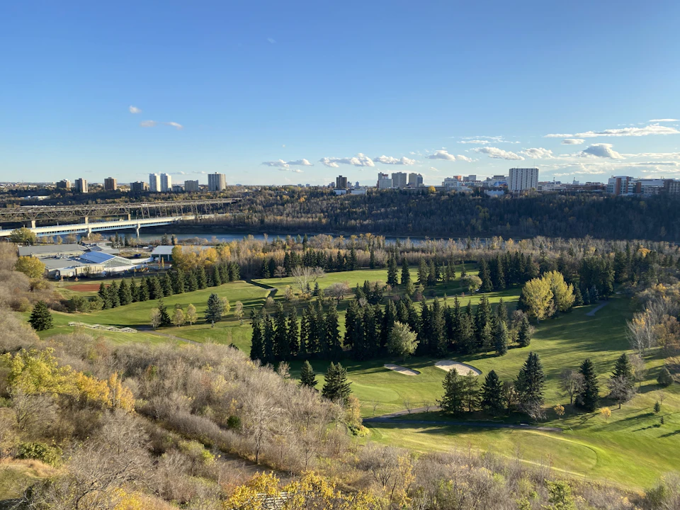 The image depicts a serene golf course nestled amidst nature, with a cityscape serving as a backdrop. The lush green fairways and trees are bathed in sunlight, creating a picturesque scene. In the distance, the city skyline is visible, adding depth to the landscape. The perspective of the photo is from above, providing an expansive view of the golf course and its surroundings.