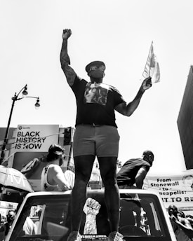 A person stands atop a vehicle, raising a fist and holding a flag, wearing a mask and glasses. The background features a billboard with 'Black History is Now' and another displaying text. Several individuals are present, and the atmosphere suggests a protest or rally.