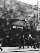 A black and white street scene with several people walking on a sidewalk in front of a brick building. The building has a sign with 'sly society' written on it and several smaller circular signs. The atmosphere is somewhat dreary with a wet surface visible, possibly indicating rain.