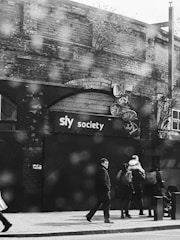 A black and white street scene with several people walking on a sidewalk in front of a brick building. The building has a sign with 'sly society' written on it and several smaller circular signs. The atmosphere is somewhat dreary with a wet surface visible, possibly indicating rain.