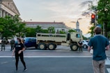 Military personnel and civilians are present at an intersection blocked by a military truck. The setting is urban, featuring historic architecture in the background. People are wearing masks and observing the scene. The surrounding area is populated with trees and street signs, suggesting a city environment.