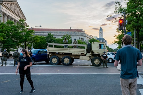 Military personnel and civilians are present at an intersection blocked by a military truck. The setting is urban, featuring historic architecture in the background. People are wearing masks and observing the scene. The surrounding area is populated with trees and street signs, suggesting a city environment.