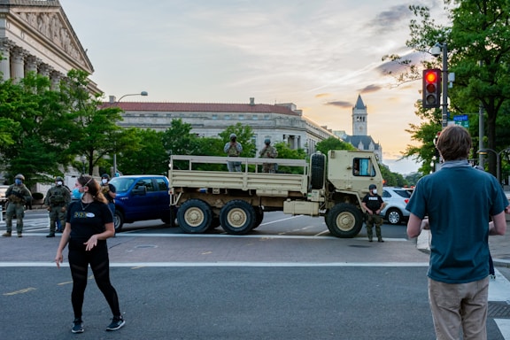 Military personnel and civilians are present at an intersection blocked by a military truck. The setting is urban, featuring historic architecture in the background. People are wearing masks and observing the scene. The surrounding area is populated with trees and street signs, suggesting a city environment.