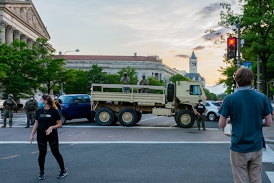 Military personnel and civilians are present at an intersection blocked by a military truck. The setting is urban, featuring historic architecture in the background. People are wearing masks and observing the scene. The surrounding area is populated with trees and street signs, suggesting a city environment.