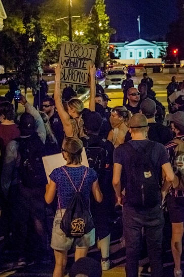 A nighttime protest scene with a crowd of people gathered, many wearing masks and holding signs. Prominently, one individual is holding a sign that reads 'Uproot White Supremacy'. The scene is illuminated by streetlights, with the White House visible in the background.