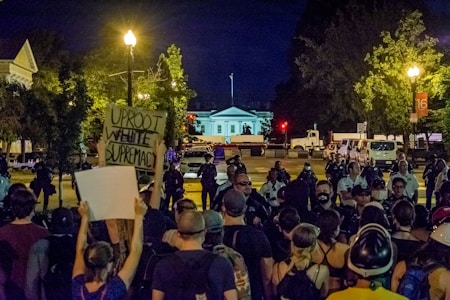 A nighttime protest with several people gathered, some holding signs. A prominent sign reads 'Uproot White Supremacy.' Police officers are seen forming a line facing the crowd. The background features a well-lit building and streetlights among trees.