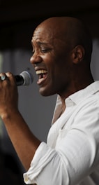 A person passionately singing into a microphone, wearing a white shirt and displaying an expressive, emotional facial expression.