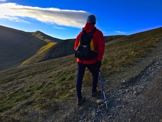 A person stands on a rugged mountain trail, equipped with hiking gear and a walking stick. The individual is wearing a red jacket, dark pants, a backpack, and a gray beanie. The landscape features rolling green hills with patches of rocky terrain and a clear blue sky overhead with a streak of white clouds.