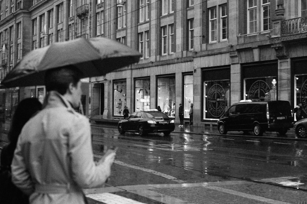grayscale photo of man in white robe holding umbrella walking on pedestrian lane