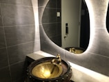A modern bathroom scene features a textured gray tiled wall and a stylish, oval backlit mirror. A unique, artistic basin resembling a bronze cauldron with a floral design is paired with an ornate faucet. Reflections can be seen in the mirror, and a metallic door handle is visible in the background.