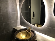 A modern bathroom scene features a textured gray tiled wall and a stylish, oval backlit mirror. A unique, artistic basin resembling a bronze cauldron with a floral design is paired with an ornate faucet. Reflections can be seen in the mirror, and a metallic door handle is visible in the background.