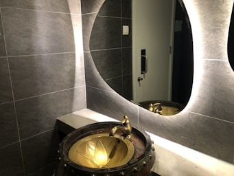 A modern bathroom scene features a textured gray tiled wall and a stylish, oval backlit mirror. A unique, artistic basin resembling a bronze cauldron with a floral design is paired with an ornate faucet. Reflections can be seen in the mirror, and a metallic door handle is visible in the background.