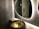 A modern bathroom scene features a textured gray tiled wall and a stylish, oval backlit mirror. A unique, artistic basin resembling a bronze cauldron with a floral design is paired with an ornate faucet. Reflections can be seen in the mirror, and a metallic door handle is visible in the background.