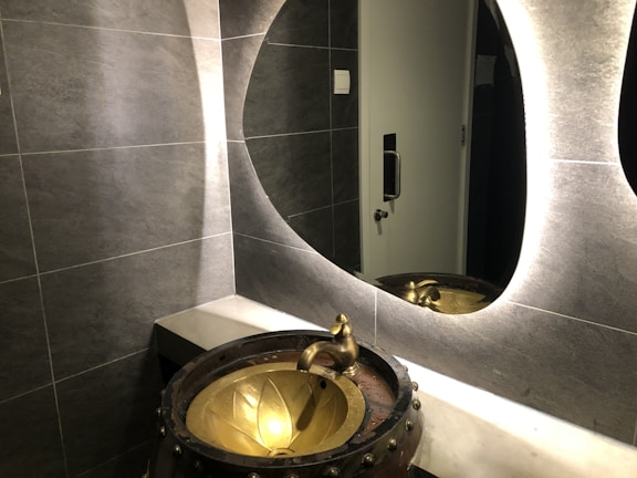 A modern bathroom scene features a textured gray tiled wall and a stylish, oval backlit mirror. A unique, artistic basin resembling a bronze cauldron with a floral design is paired with an ornate faucet. Reflections can be seen in the mirror, and a metallic door handle is visible in the background.
