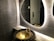 A modern bathroom scene features a textured gray tiled wall and a stylish, oval backlit mirror. A unique, artistic basin resembling a bronze cauldron with a floral design is paired with an ornate faucet. Reflections can be seen in the mirror, and a metallic door handle is visible in the background.