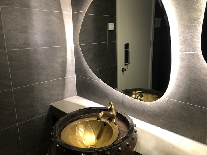A modern bathroom scene features a textured gray tiled wall and a stylish, oval backlit mirror. A unique, artistic basin resembling a bronze cauldron with a floral design is paired with an ornate faucet. Reflections can be seen in the mirror, and a metallic door handle is visible in the background.