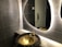 A modern bathroom scene features a textured gray tiled wall and a stylish, oval backlit mirror. A unique, artistic basin resembling a bronze cauldron with a floral design is paired with an ornate faucet. Reflections can be seen in the mirror, and a metallic door handle is visible in the background.