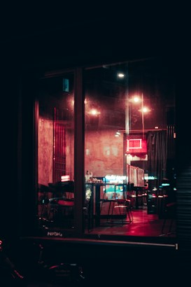Inside a dimly lit restaurant or bar, red neon lights cast a warm glow on the interior. The setting includes empty tables and chairs, and the reflection of city lights can be seen through the large glass windows.