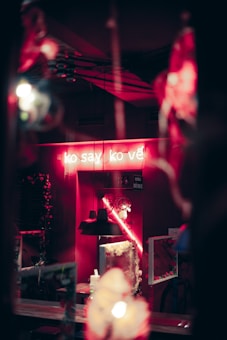 A dimly lit interior featuring a neon sign with text in a foreign language. Neon lighting casts a pinkish-red glow, illuminating various decorations and furniture like tables and hanging lights. Reflections in what appears to be a mirror add complexity to the composition.
