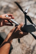 Hands holding a small drone outdoors, with focus on the propellers and body of the drone. The background is slightly blurred, suggesting an outdoor setting possibly on a sunny day.