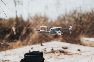 A drone hovers in mid-air in an outdoor setting with a blurred natural background. Below the drone, a black bag sits on a flat surface.