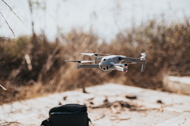 A drone hovers in mid-air in an outdoor setting with a blurred natural background. Below the drone, a black bag sits on a flat surface.