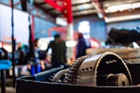 A close-up view of industrial machinery, possibly a gear mechanism, with a blurred background that includes people and additional equipment. The setting appears to be a workshop or factory environment with metal structures and vibrant lighting.