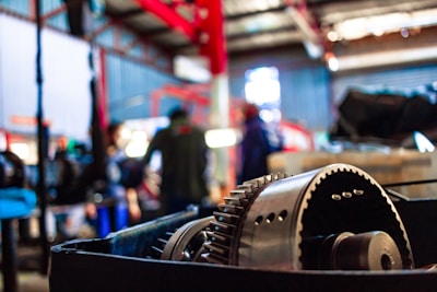 A close-up view of industrial machinery, possibly a gear mechanism, with a blurred background that includes people and additional equipment. The setting appears to be a workshop or factory environment with metal structures and vibrant lighting.