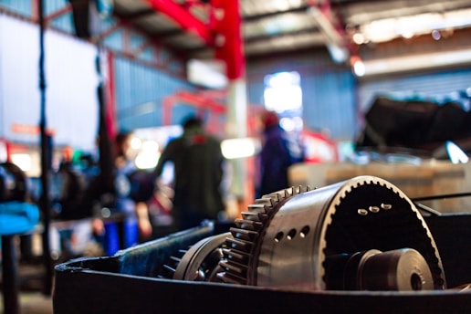 A close-up view of industrial machinery, possibly a gear mechanism, with a blurred background that includes people and additional equipment. The setting appears to be a workshop or factory environment with metal structures and vibrant lighting.