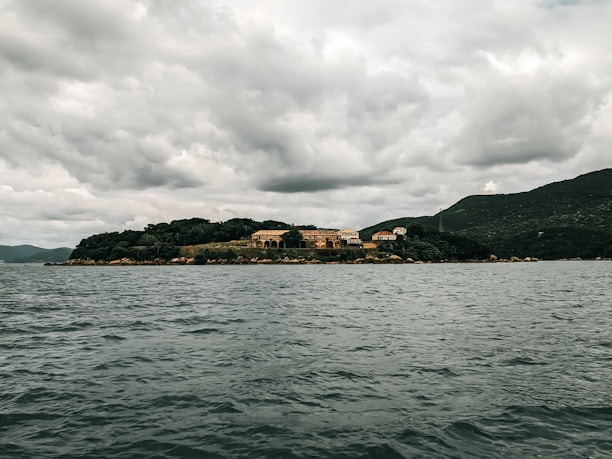 A small island features several buildings surrounded by dense greenery. The island is set against a backdrop of rolling hills under an overcast sky. The scene includes a large body of water in the foreground, with gentle ripples indicating calm conditions.