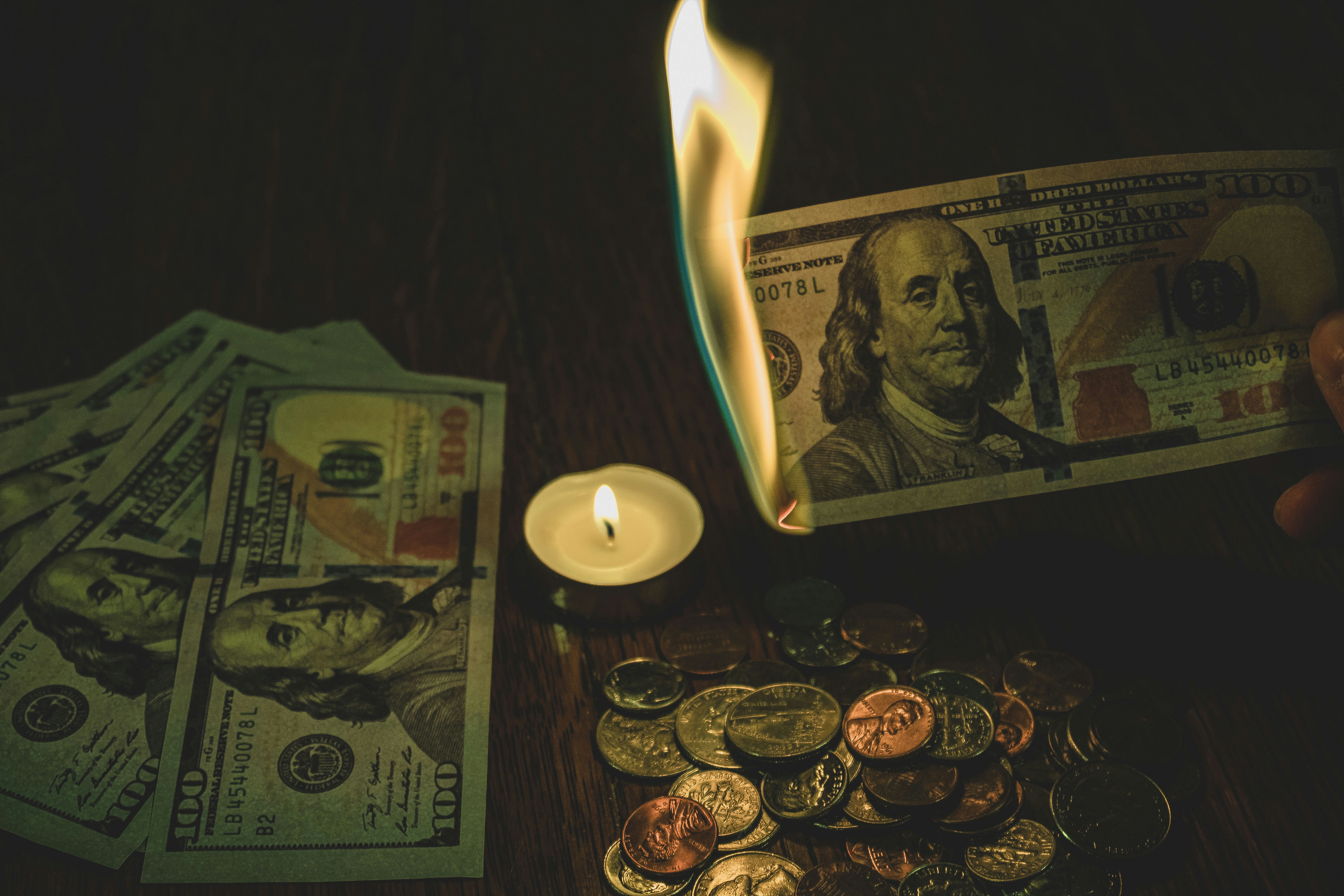 100 dollar bill in flames over candle surrounded by coins and 100 dollar bills.