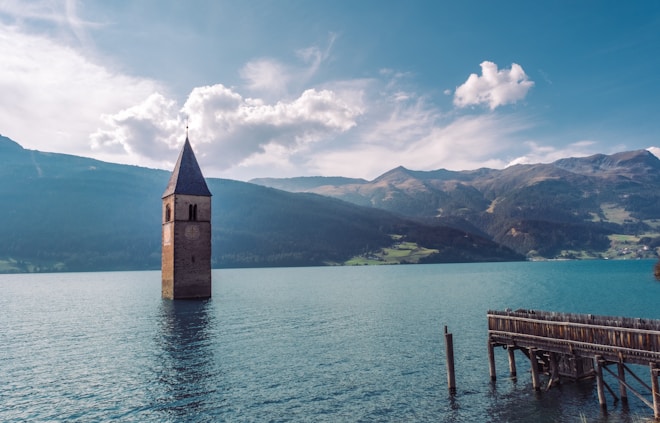 A picturesque scene featuring a submerged bell tower standing in the middle of a calm, blue lake surrounded by mountains. The sky is clear with a few clouds, and a wooden pier extends into the water on the right.