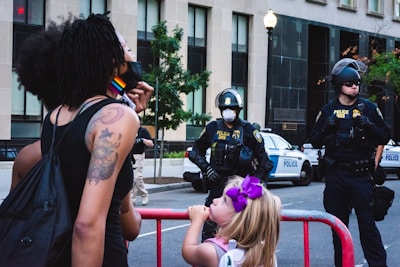 A woman with tattoos wearing a rainbow-patterned mask and a young girl with a purple bow in her hair stand near a red barricade, facing three police officers in uniform. The officers are wearing protective gear, including helmets and masks, and are positioned in front of police vehicles on a city street. In the background are buildings and trees, with a street lamp visible.