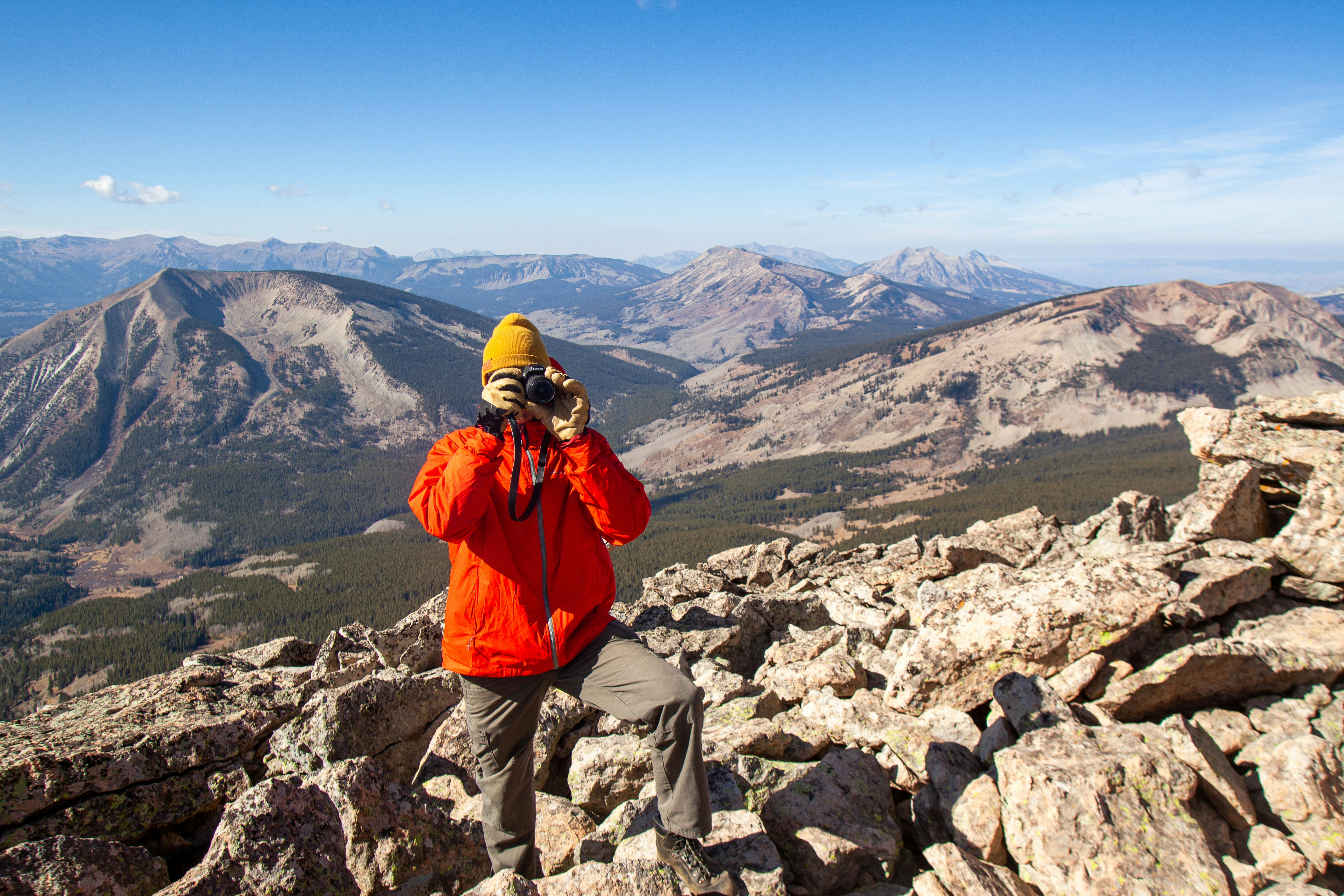 man in orange jacket and brown pants wearing yellow helmet sitting on rocky mountain during daytime