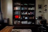 A cozy home office and reading nook featuring a large wooden bookshelf filled with a variety of books, decorative items, and plants. There's a coffee table in front, and a small desk to the right with a laptop, lamp, and a framed portrait above it. The room is softly lit, with a warm and inviting atmosphere.