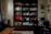 A cozy home office and reading nook featuring a large wooden bookshelf filled with a variety of books, decorative items, and plants. There's a coffee table in front, and a small desk to the right with a laptop, lamp, and a framed portrait above it. The room is softly lit, with a warm and inviting atmosphere.