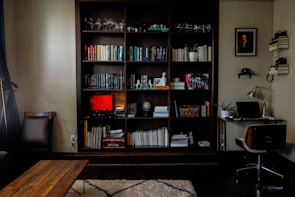 A cozy home office and reading nook featuring a large wooden bookshelf filled with a variety of books, decorative items, and plants. There's a coffee table in front, and a small desk to the right with a laptop, lamp, and a framed portrait above it. The room is softly lit, with a warm and inviting atmosphere.