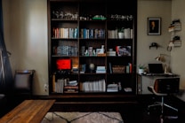 A cozy home office and reading nook featuring a large wooden bookshelf filled with a variety of books, decorative items, and plants. There's a coffee table in front, and a small desk to the right with a laptop, lamp, and a framed portrait above it. The room is softly lit, with a warm and inviting atmosphere.