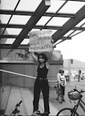 A person stands on a public staircase holding a sign with a message supporting the Black Lives Matter movement. The scene is set against a backdrop of a modern building with glass panels, bicycles, and another individual wearing a mask. The atmosphere suggests a protest or demonstration, capturing elements of solidarity and advocacy.