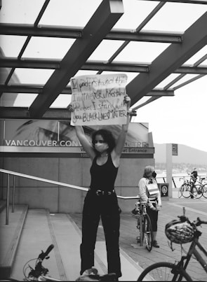 A person stands on a public staircase holding a sign with a message supporting the Black Lives Matter movement. The scene is set against a backdrop of a modern building with glass panels, bicycles, and another individual wearing a mask. The atmosphere suggests a protest or demonstration, capturing elements of solidarity and advocacy.