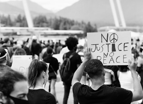A large crowd of people gathered outdoors, many wearing face masks. A prominent sign in the foreground reads 'NO JUSTICE NO PEACE' with a peace symbol replacing the letter 'O'. The background includes trees and structural elements, suggesting a public area.