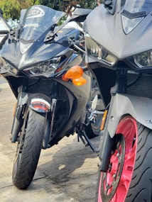 Two sleek, dark-colored sports motorcycles are parked closely together. The motorcycles feature sharp, angular designs with prominent headlights and reflective surfaces. One of the bikes has a red wheel rim, contrasting with the otherwise monochromatic color scheme.