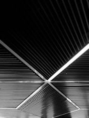 A geometric ceiling design featuring intersecting diagonal lines and rectangular sections. The overhead pattern creates a sense of depth and perspective. A linear light is integrated within one section, adding contrast to the dark material.