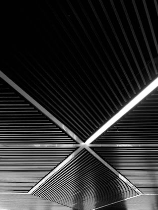 A geometric ceiling design featuring intersecting diagonal lines and rectangular sections. The overhead pattern creates a sense of depth and perspective. A linear light is integrated within one section, adding contrast to the dark material.