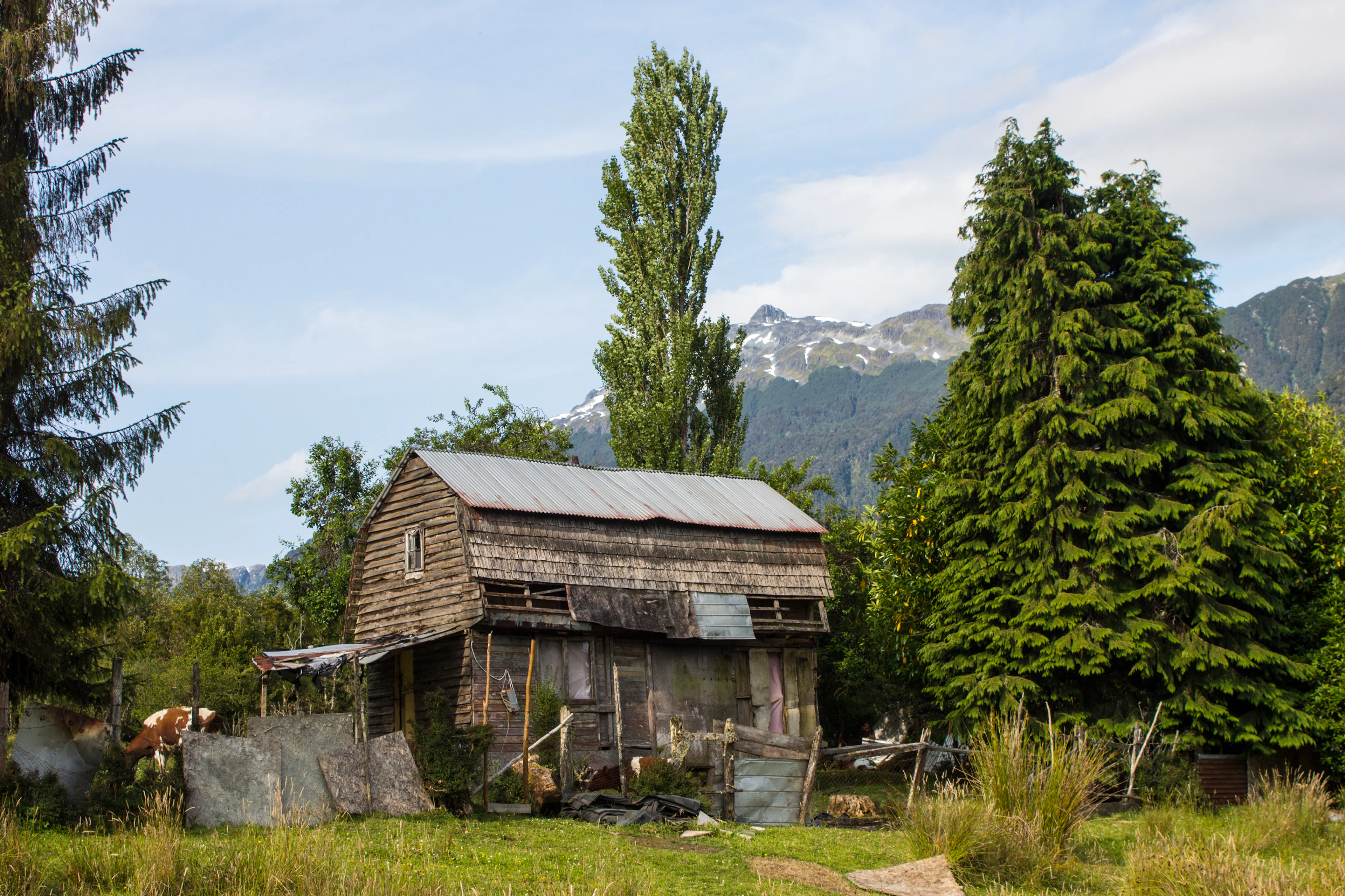 An old wooden shingle-clad house among trees with snow-capped mountains behind, Puyuhuapi, Chile
