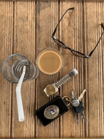 An assortment of everyday items is arranged on a wooden surface. There is a glass of iced coffee with a straw, a pair of eyeglasses, a wristwatch with a gold and silver strap, a key fob with several keys, and a leather wallet with a badge on it.