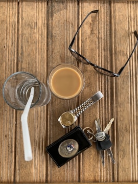 An assortment of everyday items is arranged on a wooden surface. There is a glass of iced coffee with a straw, a pair of eyeglasses, a wristwatch with a gold and silver strap, a key fob with several keys, and a leather wallet with a badge on it.