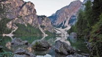 A serene mountain lake is surrounded by towering rocky cliffs and dense pine forests. The water reflects the mountains and sky, creating a tranquil and picturesque scene. Large boulders and rocks are visible in the foreground, adding texture and depth to the landscape.